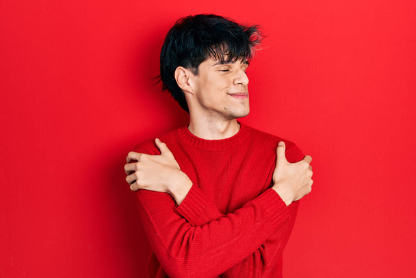 Όμορφος χίπστερ νεαρός που φοράει κόκκινο χειμωνιάτικο πουλόβερ αγκαλιάζοντας τον εαυτό του χαρούμενο και θετικό, χαμογελώντας με αυτοπεποίθηση. αυτοαγάπη και αυτοφροντίδα  - Φωτογραφία, εικόνα