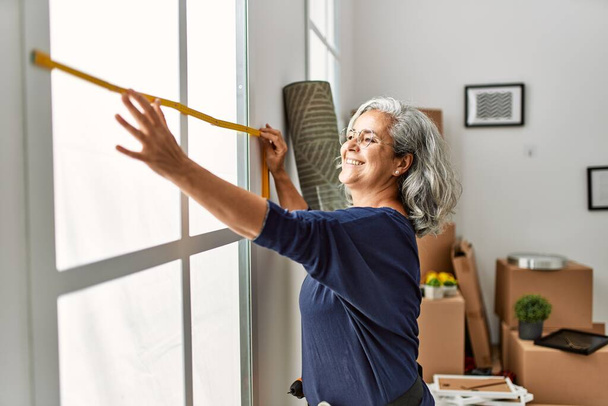 Mujer de pelo gris de mediana edad sonriendo feliz reparación de la ventana en el nuevo hogar. - Foto, imagen