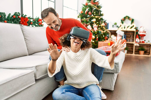 Spanisches Paar mittleren Alters lächelt glücklich auf dem Sofa sitzend und spielt zu Hause mit Virtual-Reality-Brillen. - Foto, Bild