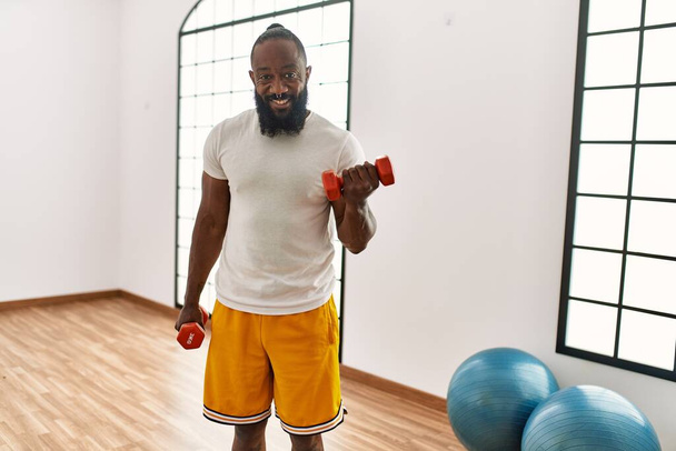 Young african american man smiling confident training with dumbbells at sport center - Photo, image