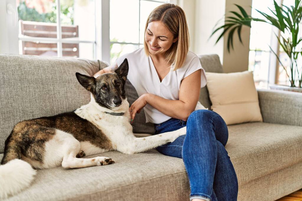 Joven chica caucásica sonriendo feliz sentado en el sofá con perro en casa. - Foto, Imagen