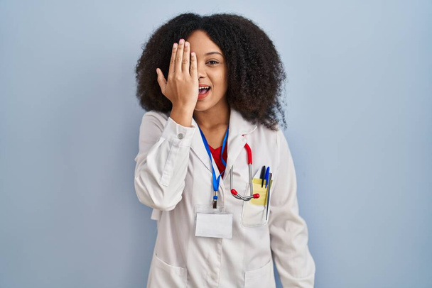 Jeune femme afro-américaine portant l'uniforme de médecin et stéthoscope couvrant un œil avec la main, sourire confiant sur le visage et l'émotion surprise.  - Photo, image