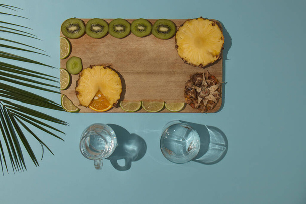 Rebanada de fruta de verano en plato de madera sobre fondo azul claro con hojas de palma y vasos con tonos claros. Minimalista plano laico escena. - Foto, imagen