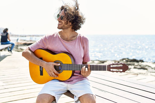Mladý hispánec hraje na klasickou kytaru sedící na lavičce na pláži. - Fotografie, Obrázek