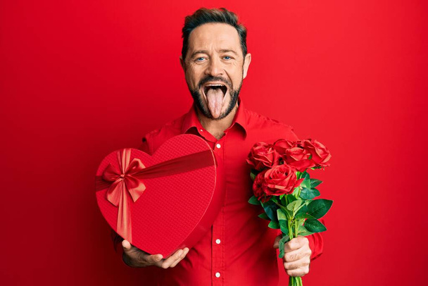 Middle age man holding anniversary present and bouquet of flowers sticking tongue out happy with funny expression.  - Photo, Image