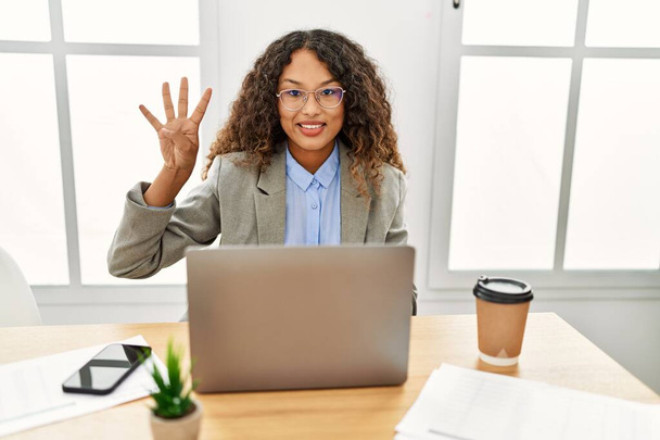 Piękna latynoska kobieta biznesu siedzi na biurku w biurze pracując z laptopem pokazując i wskazując palcami numer cztery uśmiechając się pewny siebie i szczęśliwy.  - Zdjęcie, obraz