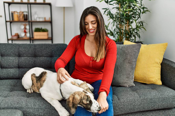 Νεαρή γυναίκα χαμογελά αυτοπεποίθηση κάθεται στον καναπέ με το σκυλί στο σπίτι - Φωτογραφία, εικόνα