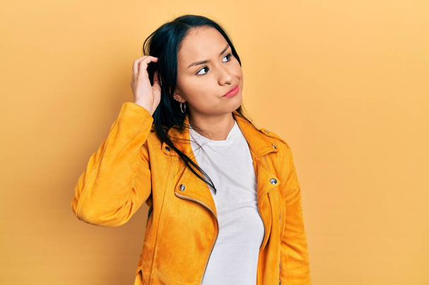 Beautiful hispanic woman with nose piercing wearing yellow leather jacket confuse and wondering about question. uncertain with doubt, thinking with hand on head. pensive concept.  - Foto, Imagen