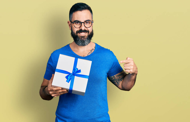 Латиноамериканець з бородою, який тримає подарунок посміхаючись щасливий вказуючи рукою і пальцем  - Фото, зображення
