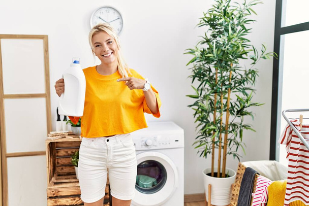Jovem caucasiana fazendo lavanderia segurando garrafa detergente sorrindo feliz apontando com a mão e o dedo  - Foto, Imagem