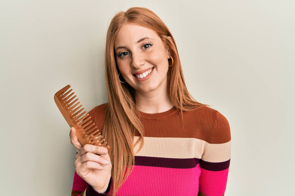 Jovem irlandês mulher styling cabelo usando pente olhando positivo e feliz de pé e sorrindo com um sorriso confiante mostrando dentes  - Foto, Imagem