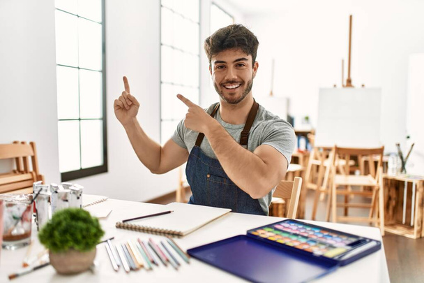 Jeune homme hispanique au studio d'art souriant et regardant la caméra pointant avec deux mains et doigts sur le côté.  - Photo, image
