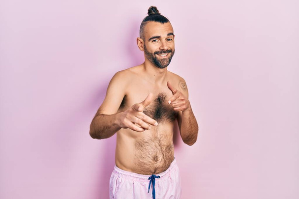 Jonge Spaanse man zonder shirt in badpak wijzend met vingers naar de camera met vrolijk en grappig gezicht. goede energie en vibes.  - Foto, afbeelding