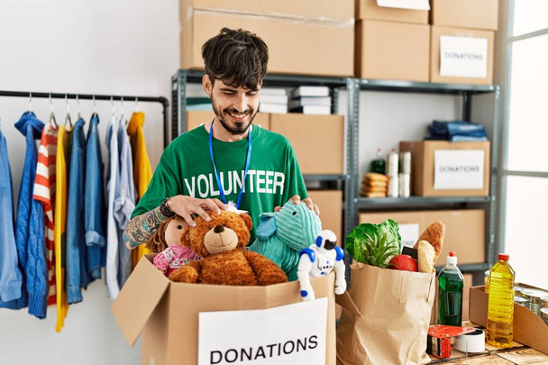 Giovane uomo ispanico che indossa uniforme volontaria con scatola di donazioni con giocattoli al centro di beneficenza. - Foto, immagini