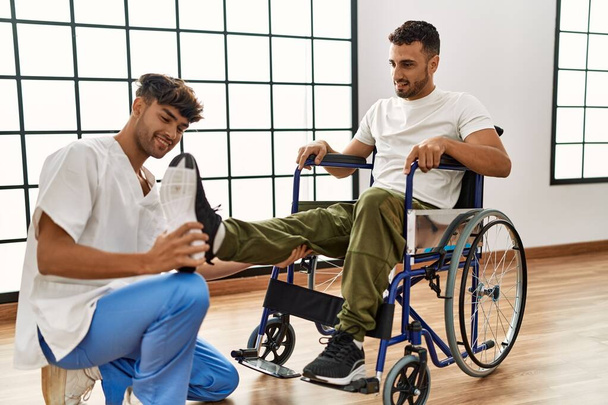 Двоє іспаномовних чоловіків фізіотерапевт і пацієнт, сидячи на інвалідному візку, мають сеанс реабілітації, що розтягує ногу в клініці
 - Фото, зображення