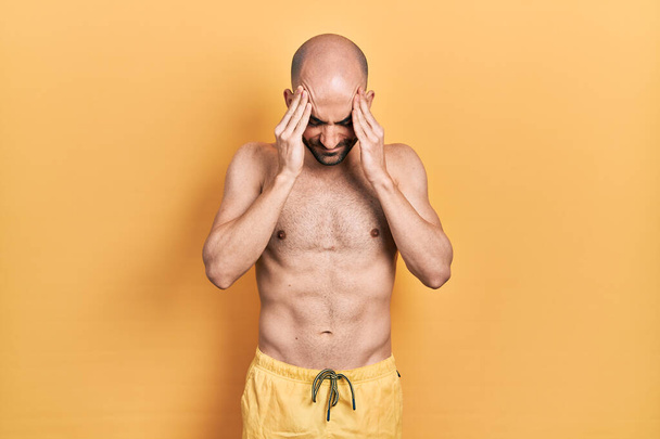 Jeune homme chauve portant des maillots de bain avec la main sur la tête pour la douleur à la tête parce que le stress. migraine souffrant.  - Photo, image