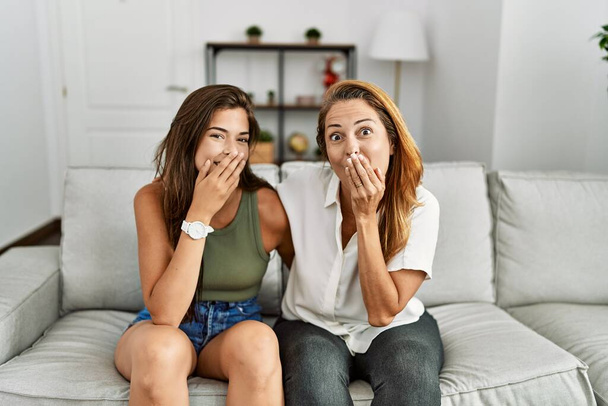 Madre e hija juntas sentadas en el sofá en casa riendo y riéndose avergonzadas cubriendo la boca con las manos, chismes y escandalosos conceptos  - Foto, imagen