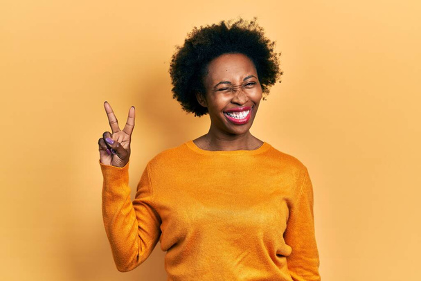 Mujer afroamericana joven vistiendo ropa casual sonriendo con la cara feliz guiñando el ojo a la cámara haciendo signo de victoria con los dedos. número dos.  - Foto, Imagen