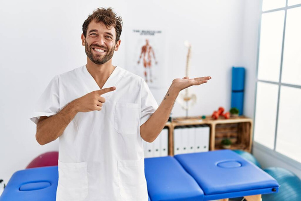 Homem fisioterapeuta bonito jovem que trabalha na clínica de recuperação da dor espantado e sorrindo para a câmera ao apresentar com a mão e apontando com o dedo.  - Foto, Imagem