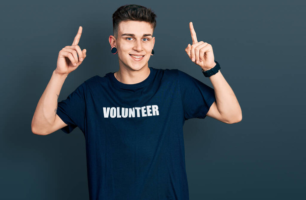 Nuori valkoihoinen poika, jolla on korvien laajentuma yllään vapaaehtoinen t-paita hymyilee hämmästynyt ja yllättynyt ja osoittaa ylös sormilla ja nosti kädet.  - Valokuva, kuva
