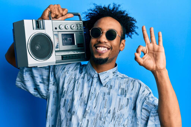 Νεαρός Αφροαμερικάνος με γενειάδα κρατώντας το κουτί, ακούγοντας μουσική κάνοντας ok υπογράψει με τα δάχτυλα, χαμογελώντας φιλικό gesturing εξαιρετικό σύμβολο  - Φωτογραφία, εικόνα