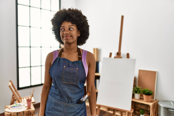 Νεαρή Αφρο-Αμερικανίδα με αφρο-μαλλιά στο στούντιο τέχνης χαμογελώντας κοιτώντας στο πλάι και κοιτάζοντας αλλού σκεπτόμενη.  - Φωτογραφία, εικόνα