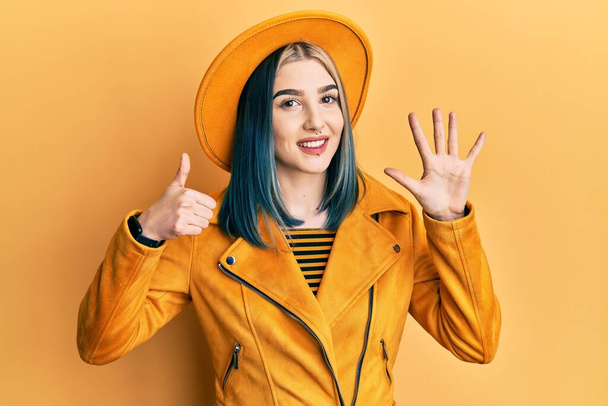 Joven chica moderna con sombrero amarillo y chaqueta de cuero mostrando y apuntando hacia arriba con los dedos número seis mientras sonríe confiado y feliz.  - Foto, Imagen
