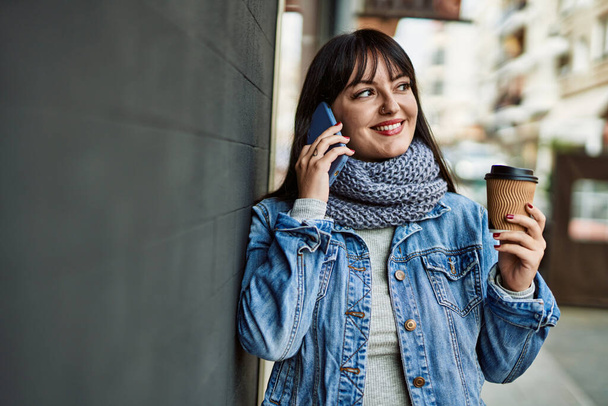Genç esmer kadın telefonda konuşuyor ve duvara yaslanmış bir fincan kahve içiyor. - Fotoğraf, Görsel