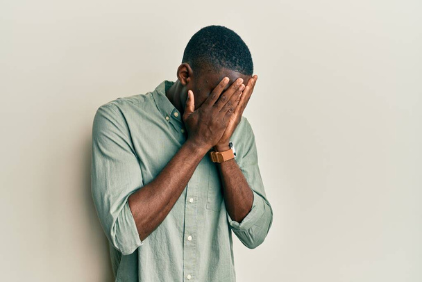 Junger afrikanisch-amerikanischer Mann in lässiger Kleidung mit traurigem Gesichtsausdruck, der das Gesicht mit Händen bedeckt, während er weint. Depressionskonzept.  - Foto, Bild