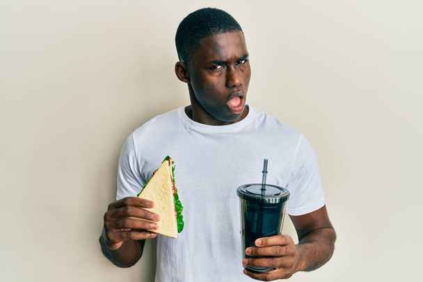 Fiatal afro-amerikai férfi eszik szendvicset és üdítőt iszik sokkos arccal, szkeptikusan és szarkasztikusan néz ki, meglepve nyitott szájjal  - Fotó, kép