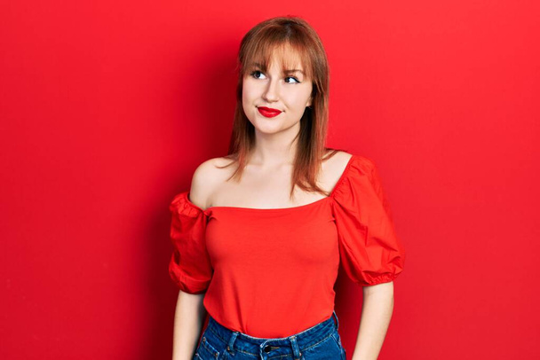 roodharige jonge vrouw dragen casual rood t shirt glimlachen kijken naar de zijkant en staren weg denken.  - Foto, afbeelding