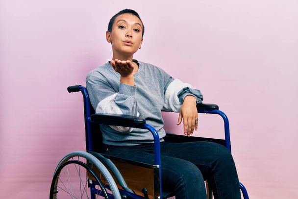 Krásná hispánská žena s krátkými vlasy sedí na invalidním vozíku a dívá se do kamery, která posílá polibek s rukou na vzduchu, je krásná a sexy. výraz lásky.  - Fotografie, Obrázek