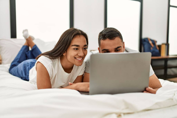 Νεαρό ζευγάρι Λατίνων χαμογελά χαρούμενος χρησιμοποιώντας φορητό υπολογιστή που βρίσκεται στο κρεβάτι. - Φωτογραφία, εικόνα
