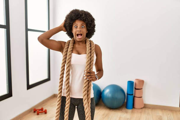 Молода афроамериканська жінка з волоссям африканського кольору під час тренувань у спортзалі з бойовими мотузками божевільна і налякана руками на голову, налякана і здивована від шоку відкритим ротом.  - Фото, зображення
