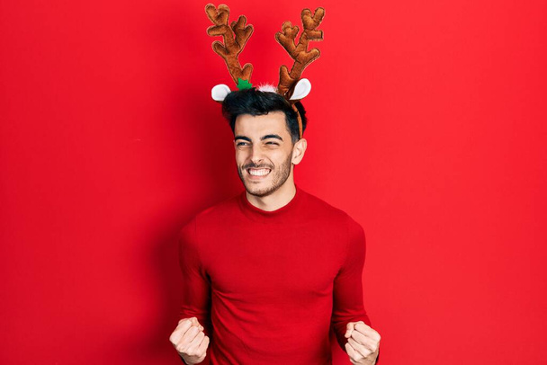 Young hispanic man wearing cute christmas reindeer horns excited for success with arms raised and eyes closed celebrating victory smiling. winner concept.  - Photo, Image