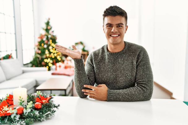 Junger hispanischer Mann sitzt auf dem Tisch am Weihnachtsbaum und lächelt fröhlich und zeigt mit der Handfläche in die Kamera.  - Foto, Bild
