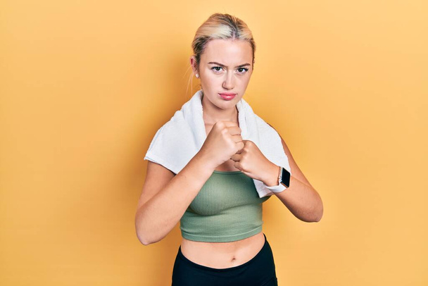 Young blonde girl wearing sportswear and towel ready to fight with fist defense gesture, angry and upset face, afraid of problem  - Foto, imagen