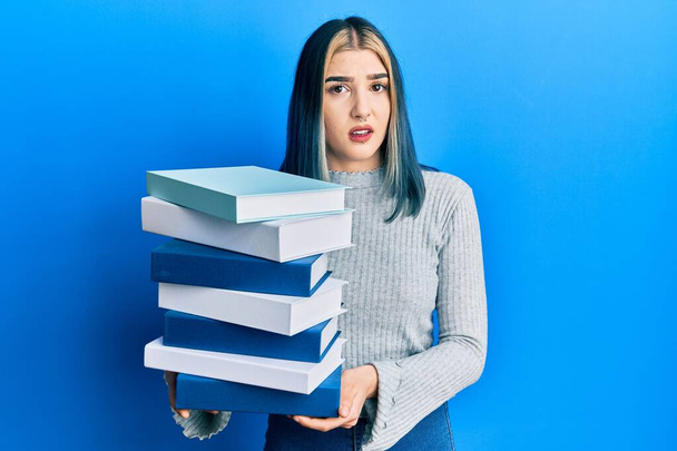 Młoda nowoczesna dziewczyna trzymająca stertę książek bez pojęcia i zdezorientowana ekspresja. koncepcja wątpliwości.  - Zdjęcie, obraz
