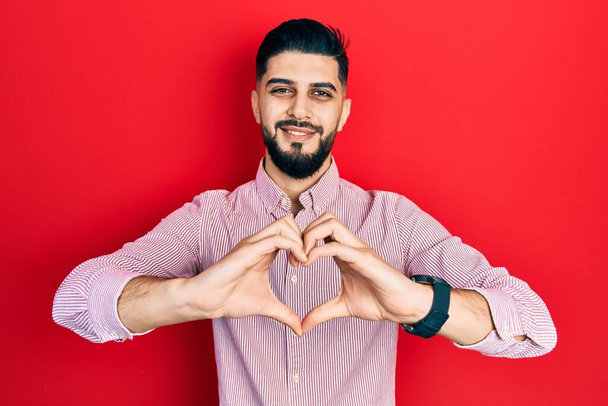 Beau homme barbu portant une chemise rouge décontractée souriant en amour montrant le symbole du cœur et la forme avec les mains. concept romantique.  - Photo, image