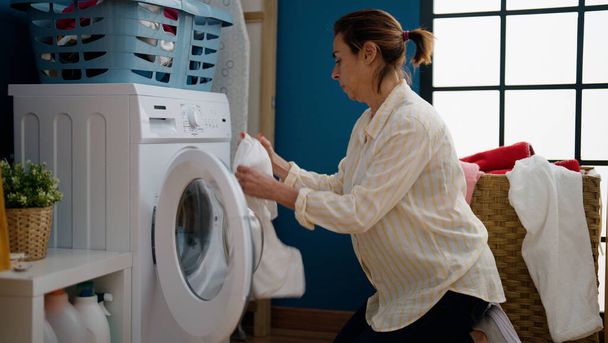 Middelbare leeftijd Spaanse vrouw wassen van kleding in wasruimte - Foto, afbeelding