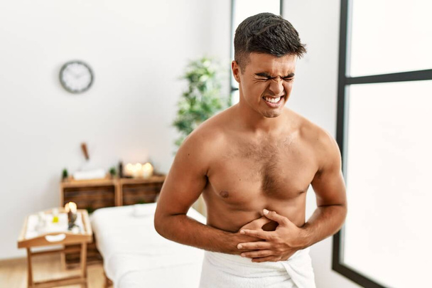 Young hispanic man standing shirtless at spa center with hand on stomach because indigestion, painful illness feeling unwell. ache concept.  - Foto, Bild