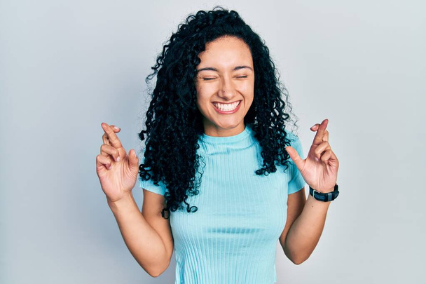 Fiatal spanyol nő göndör haj visel alkalmi kék póló gesztikuláló ujj keresztbe mosolyogva a remény és a szem csukva. szerencse és babonás fogalom.  - Fotó, kép
