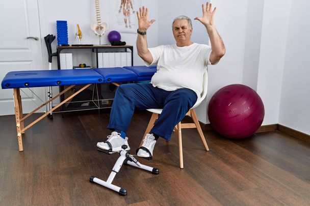 Uomo anziano caucasico presso la clinica di fisioterapia utilizzando l'esercizio a pedali mostrando e indicando con le dita numero dieci, sorridendo fiducioso e felice.  - Foto, immagini