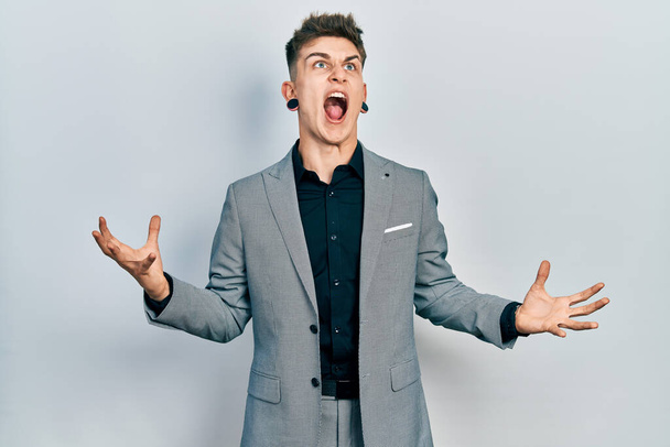 Young caucasian boy with ears dilation wearing business jacket crazy and mad shouting and yelling with aggressive expression and arms raised. frustration concept.  - Photo, Image