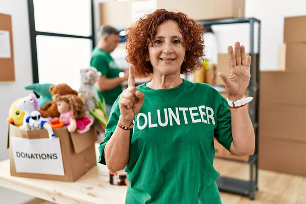 Middle age woman wearing volunteer t shirt at donations stand showing and pointing up with fingers number six while smiling confident and happy.  - Photo, Image