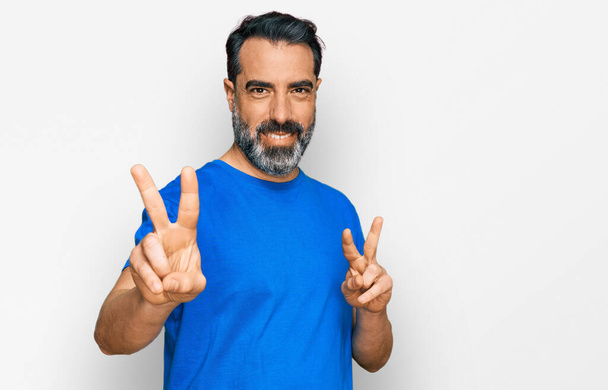Mann mittleren Alters mit Bart und lässig blauem T-Shirt lächelt in die Kamera und zeigt Finger, die Siegeszeichen machen. Nummer zwei.  - Foto, Bild