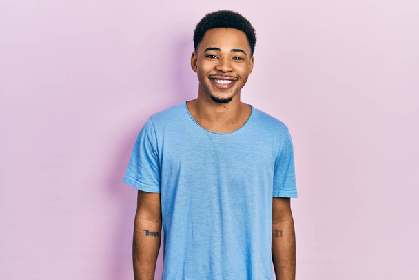 Молодой африканский американец в обычной синей футболке со счастливой и прохладной улыбкой на лице. счастливчик.  - Фото, изображение