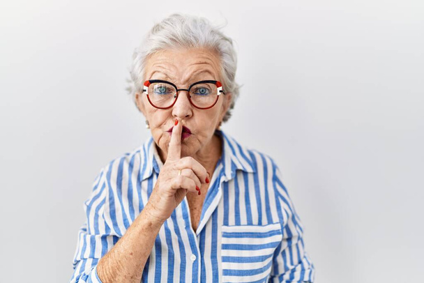 Starsza kobieta z siwymi włosami stojąca na białym tle, prosząca o ciszę z palcem na ustach. cisza i tajne pojęcie.  - Zdjęcie, obraz