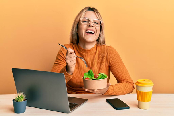 Jeune femme caucasienne travaillant au bureau mangeant une salade saine souriant et riant fort à haute voix parce que drôle blague folle.  - Photo, image