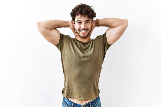 Hispanic man standing over isolated white background relaxing and stretching, arms and hands behind head and neck smiling happy  - Foto, Imagen